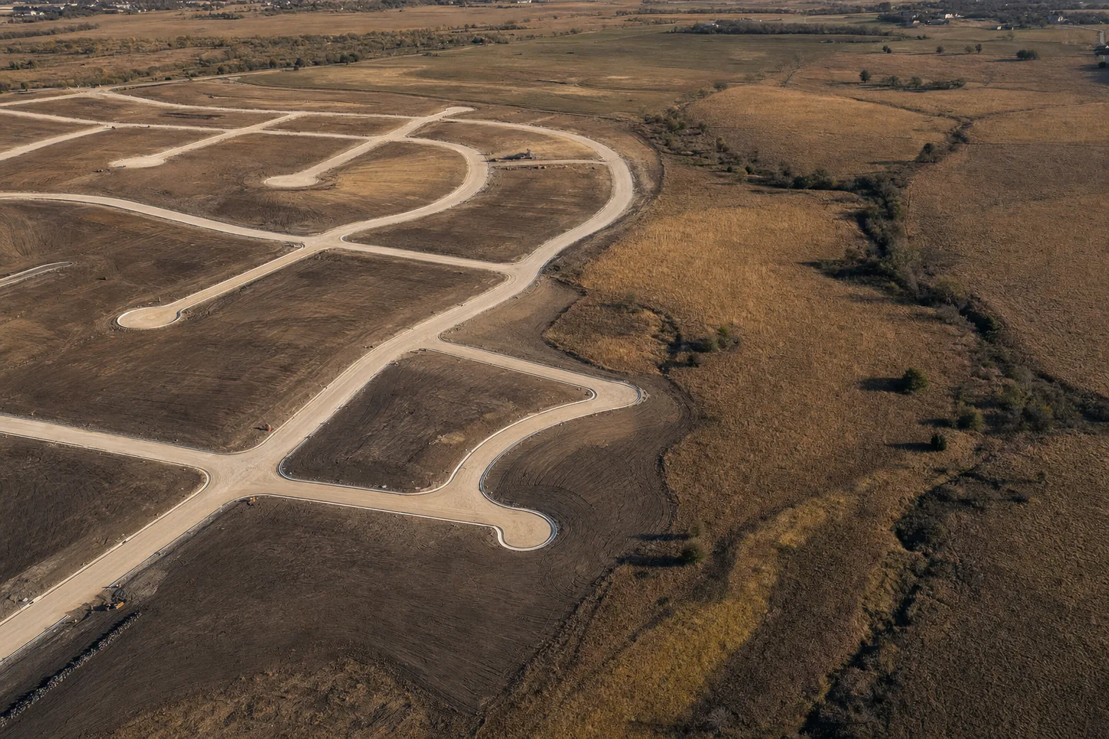 Aerial view of a newly graded subdivision with pale limestone roadbeds meeting untouched golden farmland